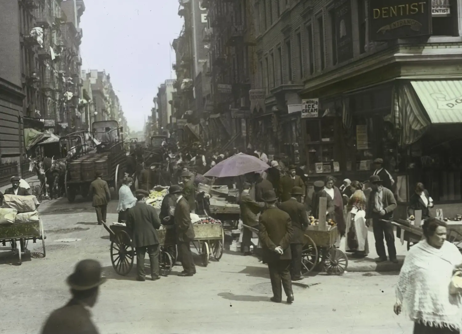 A bustling street scene. People and horse-drawn wagons crowd the road.